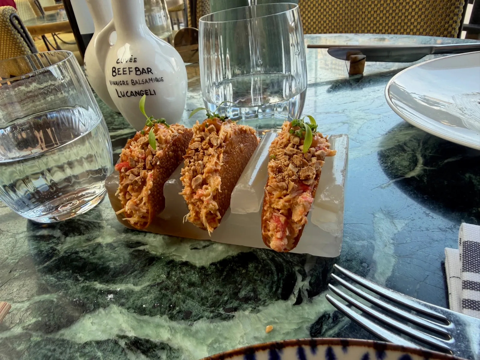 Three crispy king crab taquitos in a white marble holder at Beefbar Monte-Carlo, filled with king crab, topped with chopped cashew nuts and micro herbs, beside a branded Cuvée Beefbar balsamic bottle