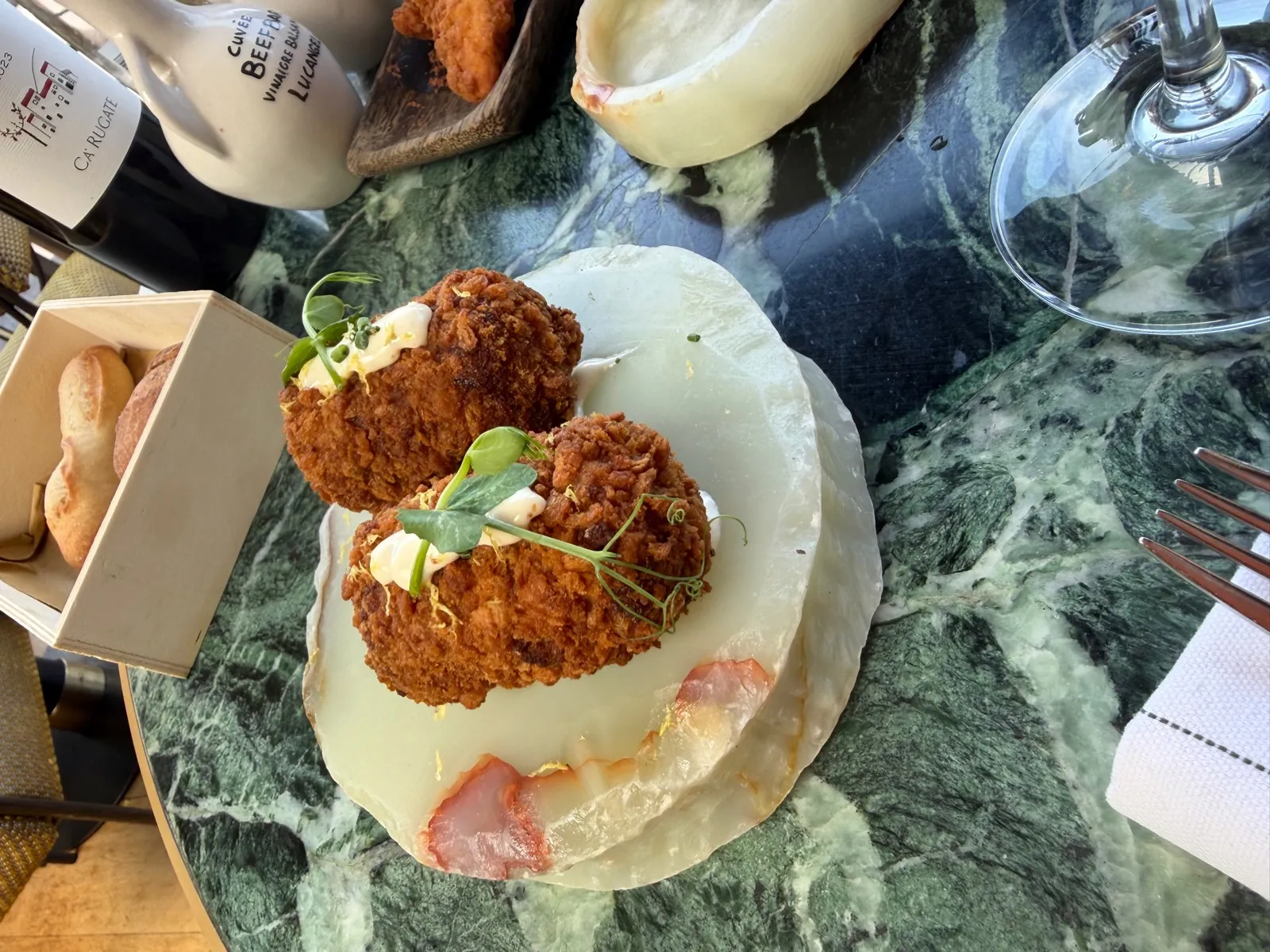 Two large golden croquettes at Beefbar Monte-Carlo topped with mayo, pea shoots and lemon zest, on a pale marble plate, with a bread box and a bottle of Ca'Rugate wine beside