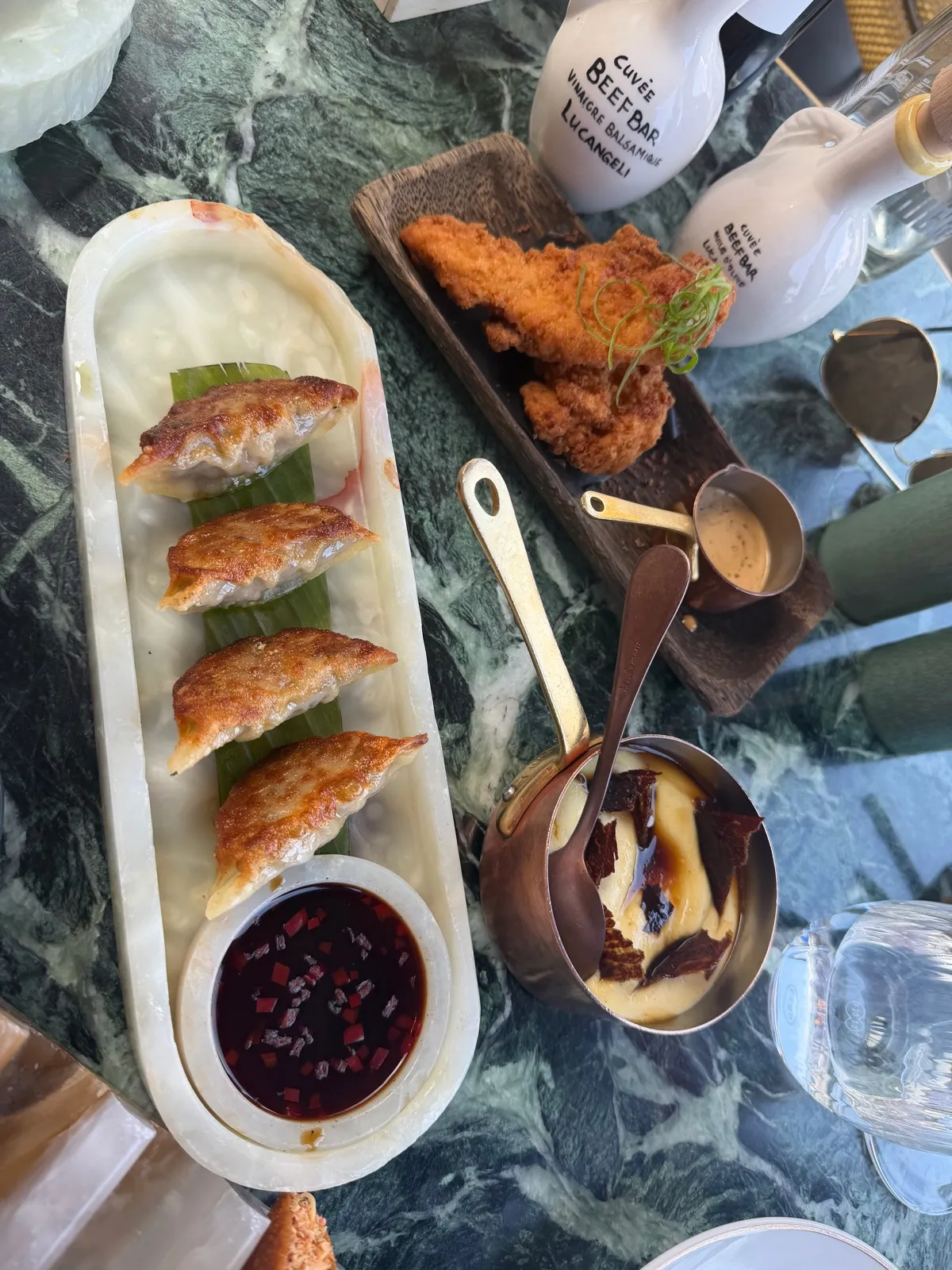 Four gyozas on a banana leaf in a marble dish at Beefbar Monte-Carlo, with a dark chilli dipping sauce, beside fried chicken on a wooden board with a gold jug of sauce