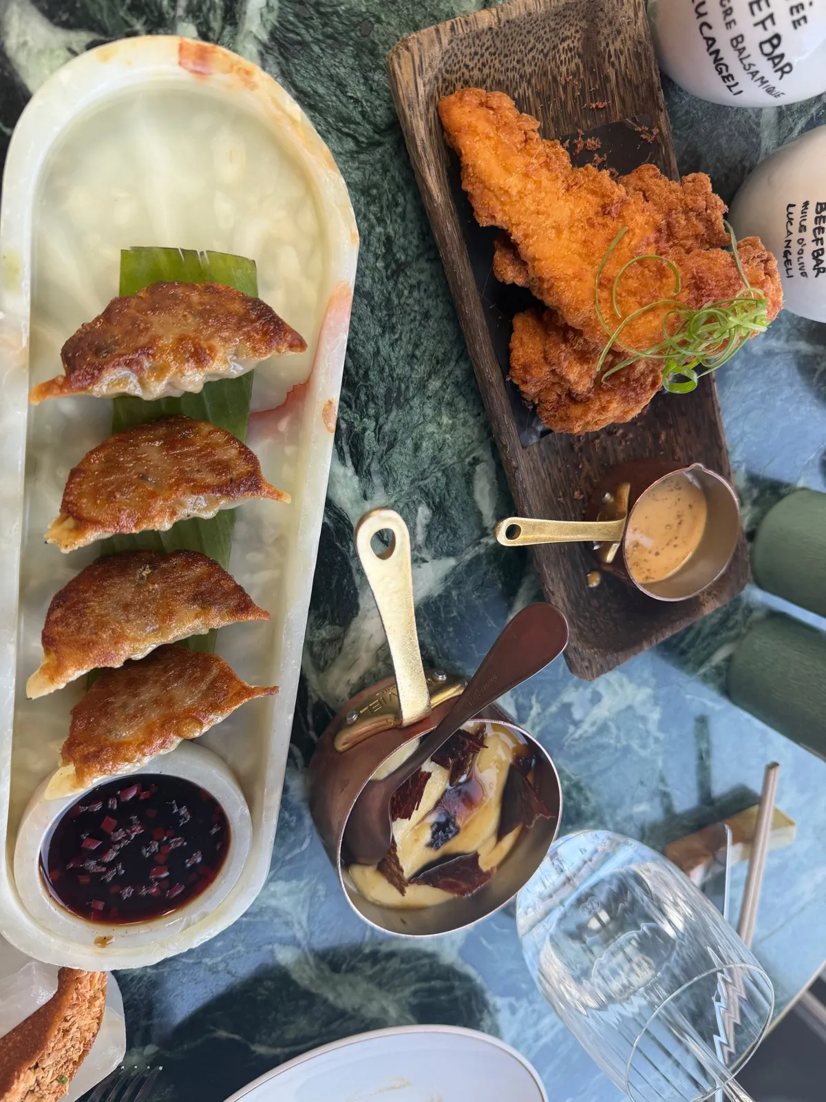 Close up of four gyozas with crisp golden bases at Beefbar Monte-Carlo, with dark chilli dipping sauce, fried chicken on a wooden board and a gold jug of pale sauce