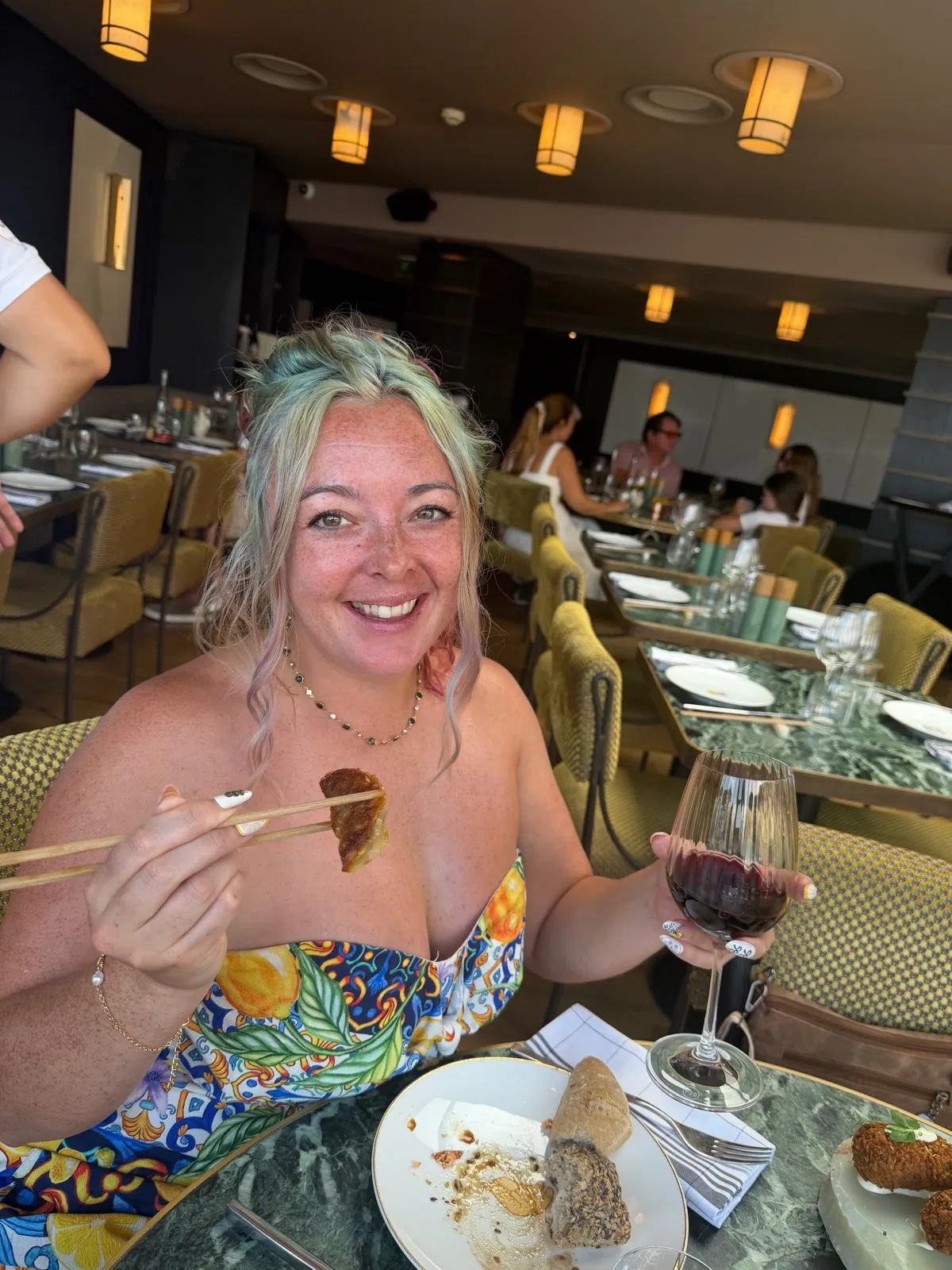 Sofi at the table at Beefbar Monte-Carlo holding a gyoza with chopsticks in one hand and a glass of red wine in the other, smiling, restaurant interior behind