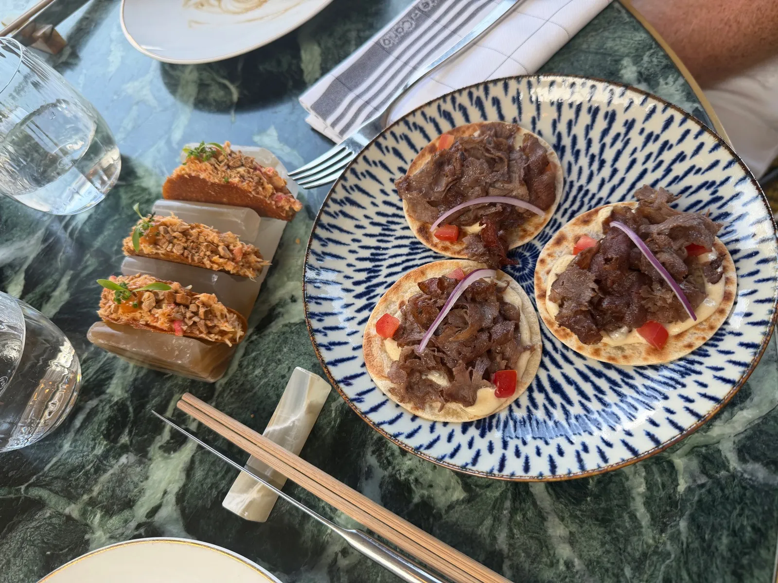 Lunch at Beefbar Monte-Carlo: a blue and white plate of three Wagyu kebabs beside three crispy king crab taquitos in a marble holder, on a green marble table
