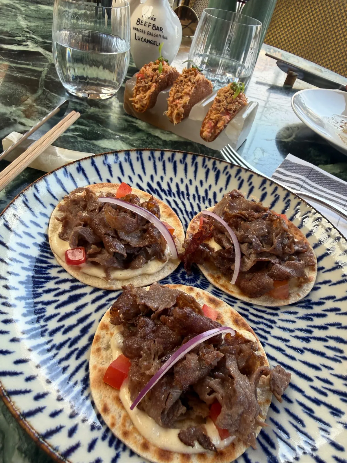 Three Wagyu kebabs at Beefbar Monte-Carlo on a blue and white patterned plate, mini Wagyu shawarma on wheat galettes with tahini, red onion and diced tomato, with three crispy king crab taquitos in a marble holder behind