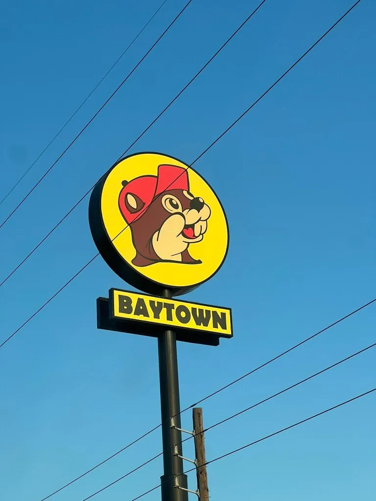 The Buc-ee's sign at the Baytown store, the beaver's head in its red cap on a yellow circle above a yellow panel reading BAYTOWN, against a bright blue sky crossed by power lines