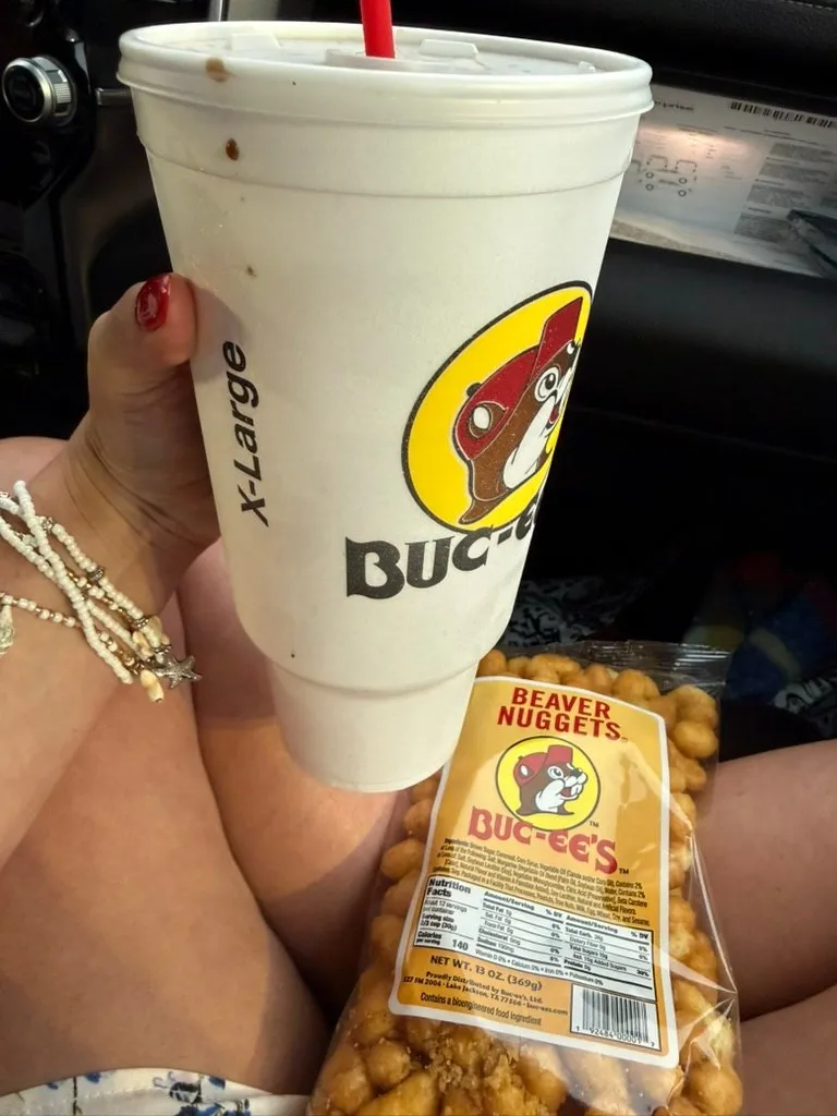 An X-Large Buc-ee's cup held up in a car, straw in the lid, next to an open bag of Beaver Nuggets on my lap