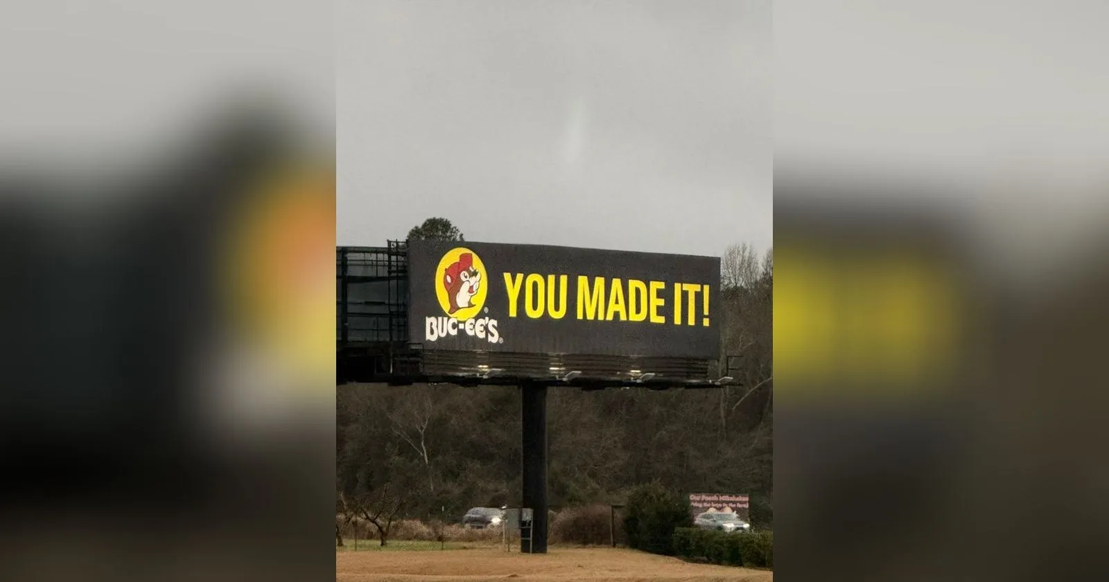 A Buc-ee's billboard beside the highway under a flat grey sky, the beaver logo in its red cap next to the words YOU MADE IT in huge yellow capitals