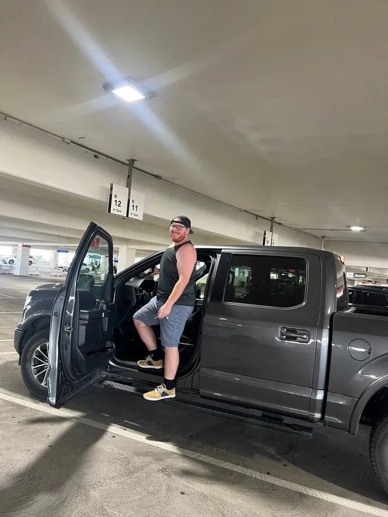 Alex stepping up into a grey Ford pickup truck in a multi-storey car park, bay markers K11 and K12 on the pillar behind him