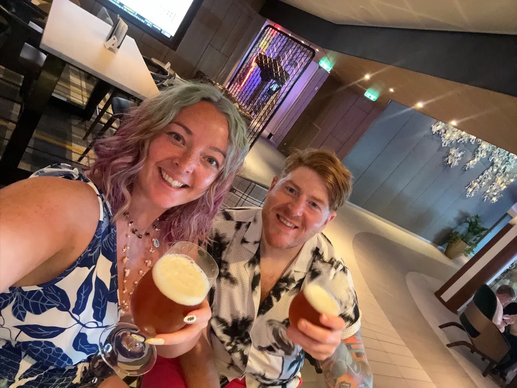 Sofi and Alex on a Celebrity ship holding up two stemmed glasses of beer with foam heads, in a restaurant with wooden ceilings and white blossom decorations behind them