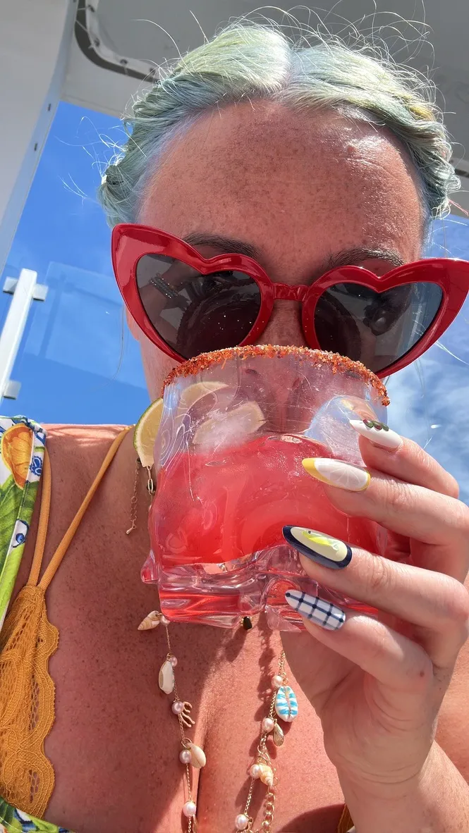Sofi in red heart-shaped sunglasses drinking a pink cocktail with a chilli salt rim and lemon slices, wearing a shell necklace, on the deck of a Celebrity ship
