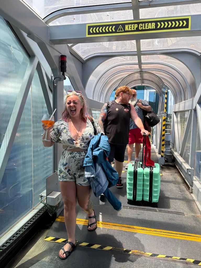 Sofi walking down the covered gangway off the ship, laughing, holding an Aperol Spritz, with Alex behind her wheeling two mint green suitcases