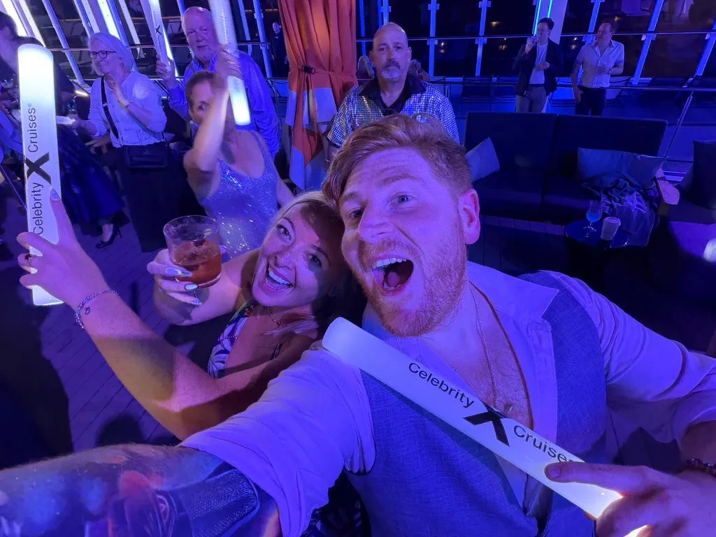 Sofi and Alex at a night deck party on a Celebrity ship holding white Celebrity Cruises glow sticks, with other guests waving glow sticks behind them