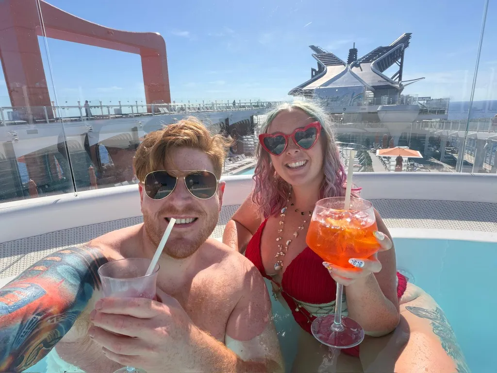 Sofi and Alex in a hot tub on the deck of a Celebrity ship, Alex with a clear drink and Sofi in a red bikini holding an Aperol Spritz, with the orange Magic Carpet platform behind them