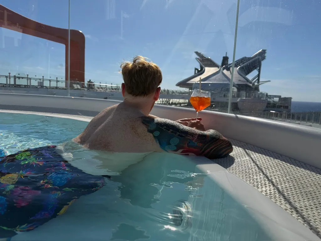 A cantilevered hot tub on the side deck of a Celebrity ship, Alex sitting in it from behind holding an Aperol Spritz, looking out towards the orange Magic Carpet platform and the sea