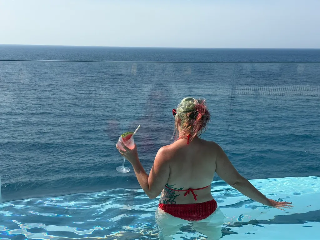 Sofi from behind in an infinity pool on a Celebrity ship, holding a pink cocktail garnished with watermelon, looking out at the sea