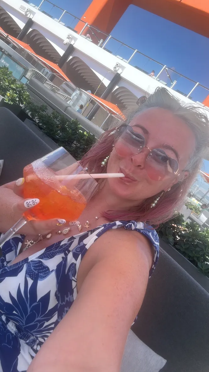 Sofi lying on a sun lounger on the deck of a Celebrity ship, wearing round pink-tinted sunglasses and pearl earrings, holding a tall Aperol Spritz up to the camera