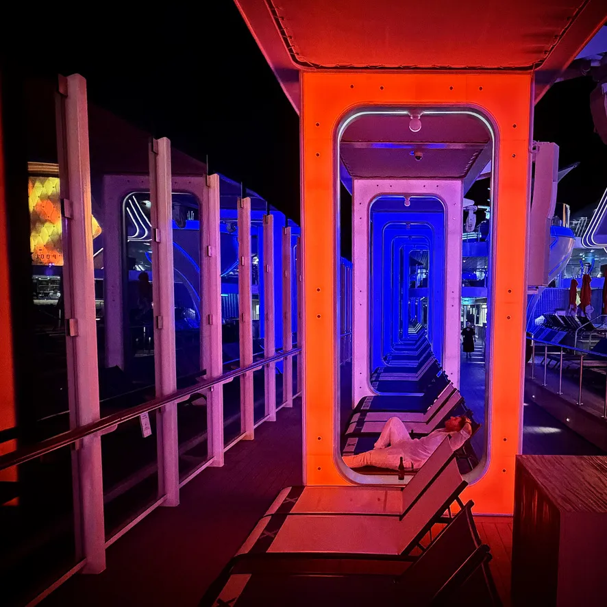 A dark deck on a Celebrity ship at night, lit by an orange doorway frame with a receding line of blue lit archways behind it and sun loungers along the side