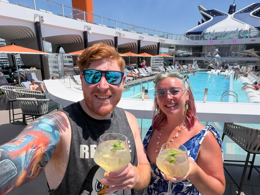 Sofi and Alex at a pool bar on a Celebrity ship holding two elderflower spritzes garnished with mint, Alex in a grey tank top and Sofi in a blue and white patterned dress
