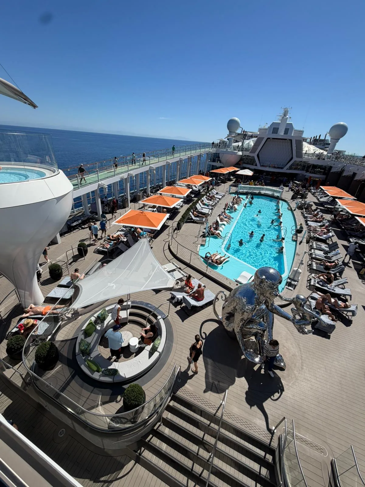 Overhead view of the pool deck on a Celebrity ship, showing a large main pool, rows of sun loungers with orange umbrellas, a chrome monkey sculpture and a hot tub cantilevered over the side, with the sea beyond