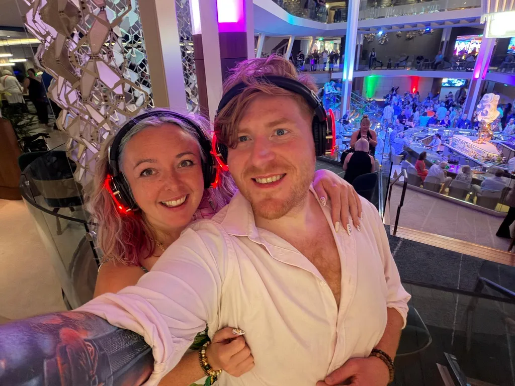 Sofi and Alex wearing light up silent disco headphones on a balcony overlooking the atrium of a Celebrity ship, with coloured lights and a crowd below
