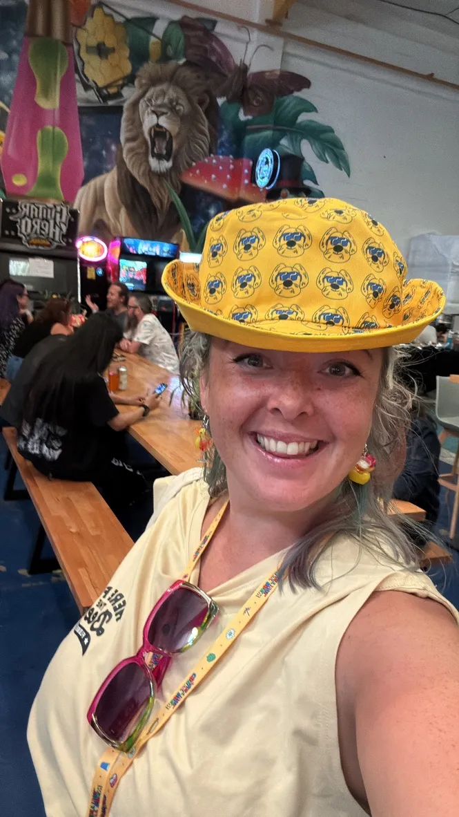 Me in a yellow bucket hat covered in cartoon dogs, won at the higher or lower card game, arcade machines and a painted lion mural behind