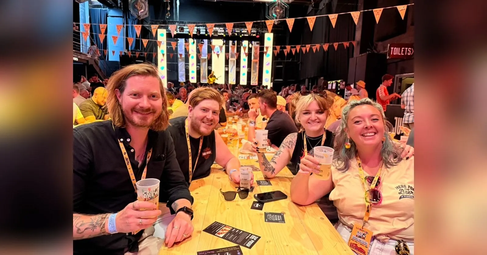 The four of us at a long wooden bench at Craft Brew Festival Liverpool, half pint cups in hand, bunting and a live act on stage behind us