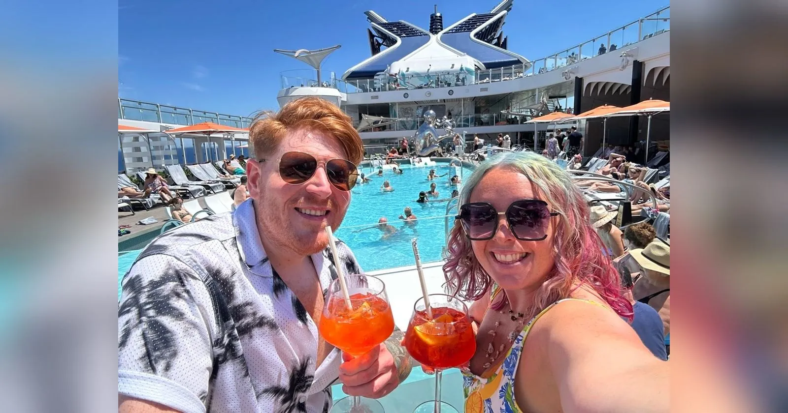 Sofi and Alex at the pool bar on a Celebrity ship, both holding tall Aperol Spritz glasses with straws, with the busy pool and the ship's funnel behind them