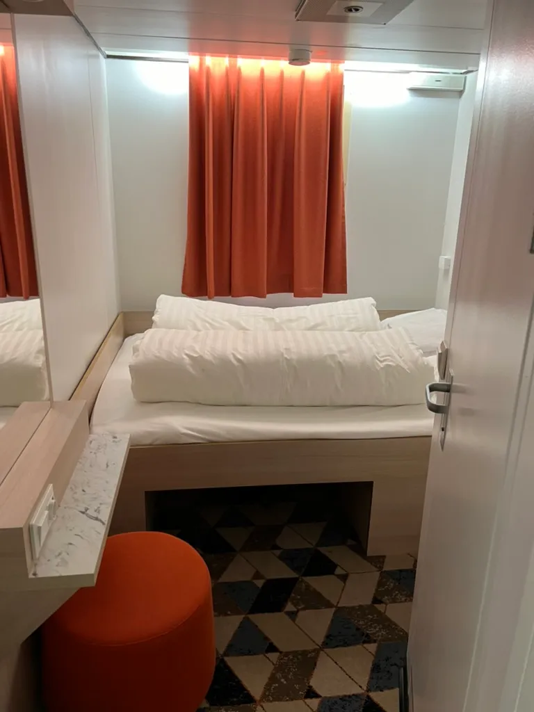 Our cabin on board: a made-up double bed with white bedding against a wooden wall, orange curtains and reading lights above it, and an orange stool at a small dressing table.