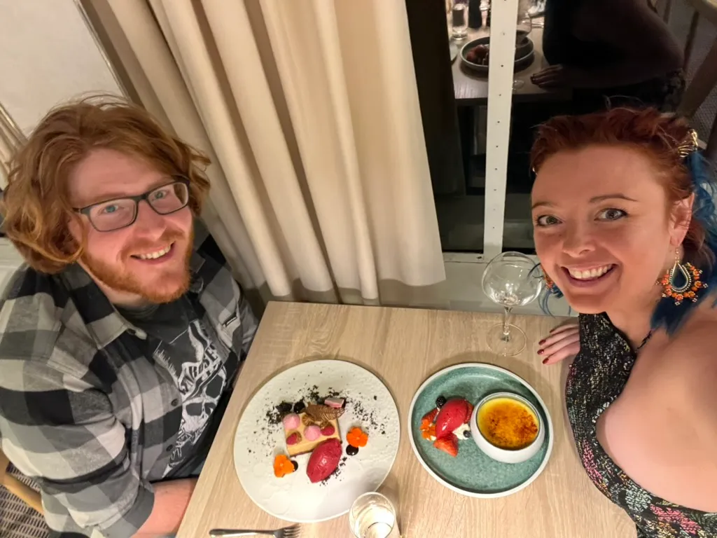 The two of us at the table in the ship's restaurant, looking up at the camera over two desserts: a tart with sorbet and crumb on a white plate, and a creme brulee with berries and sorbet on a teal plate.