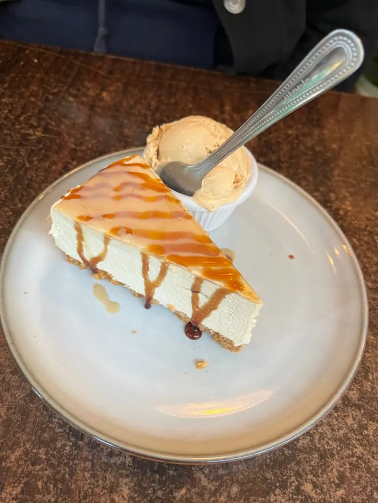 A slice of Baileys cheesecake with caramel sauce drizzled over it and a scoop of ice cream in a ramekin alongside