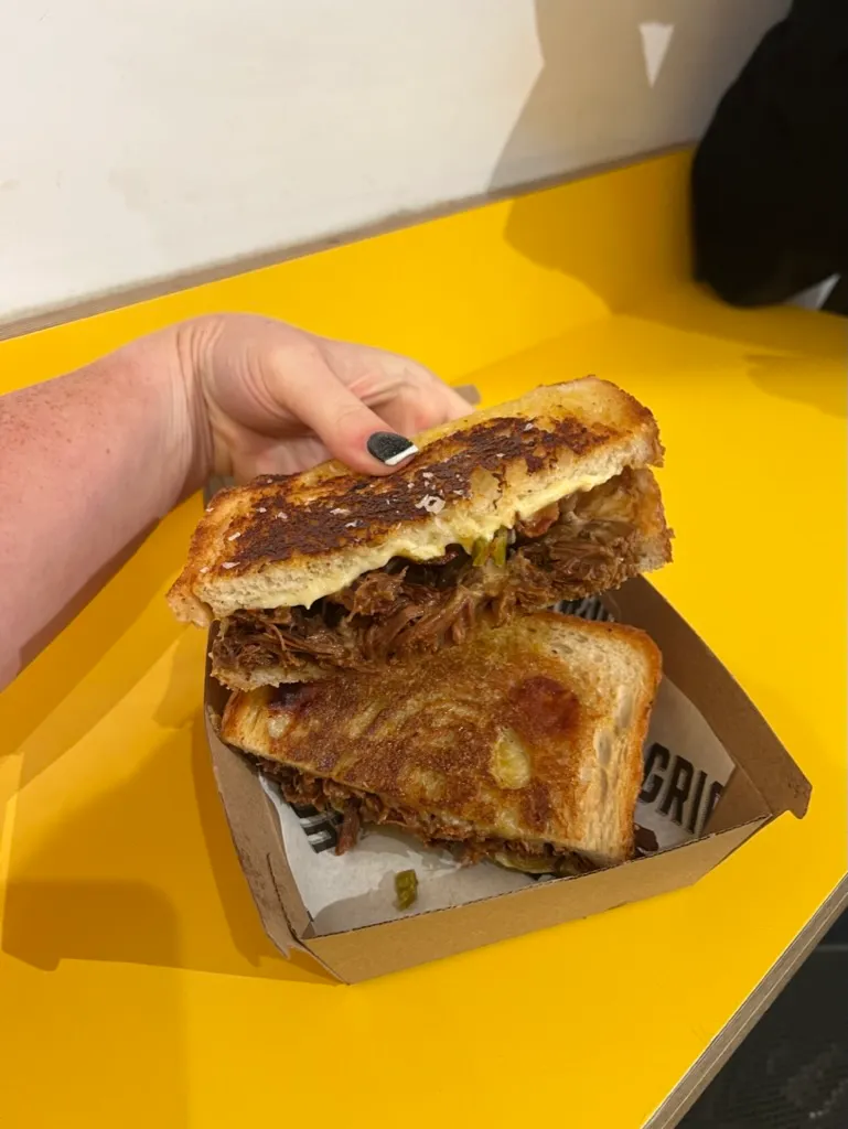 A toasted cheese and brisket sambo from Griolladh on a yellow table, one half held up to show the brisket and melted cheese inside, the other still in its cardboard box, cheese crisped over the edges of the bread