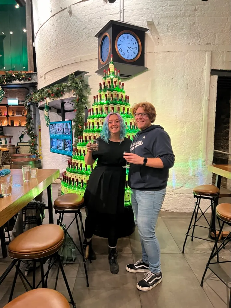 The two of us in front of a Christmas tree built entirely out of green Jameson bottles, under a black clock tower inside the distillery