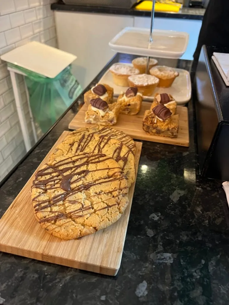 A counter of sweet things: a giant chocolate drizzled cookie on a board, tray bakes topped with Kinder bars, and mince pies on a cake stand