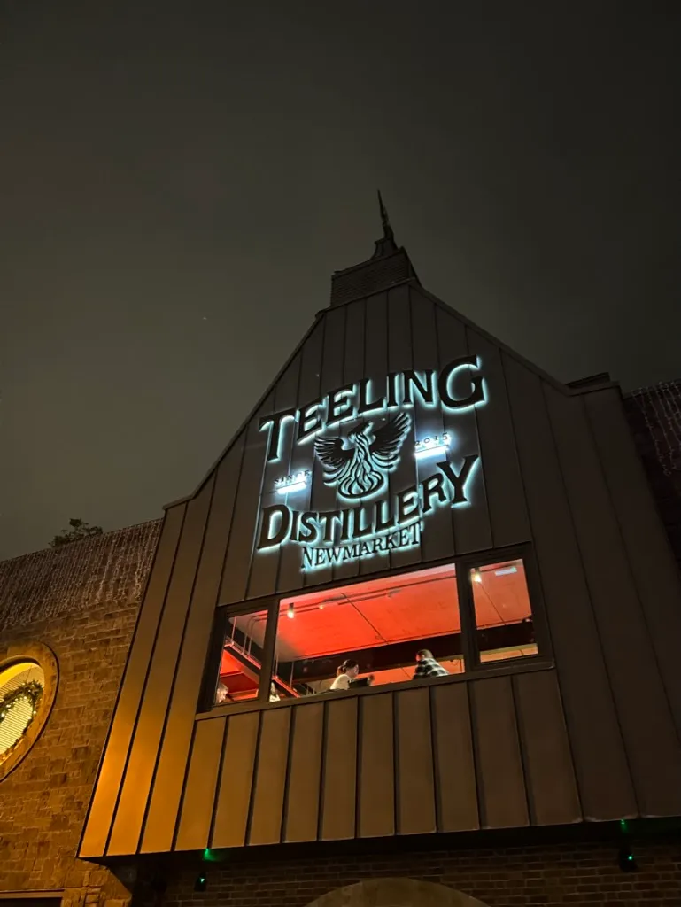 The lit Teeling Distillery sign on the dark metal gable end of the Newmarket building at night, with the phoenix logo glowing between the words