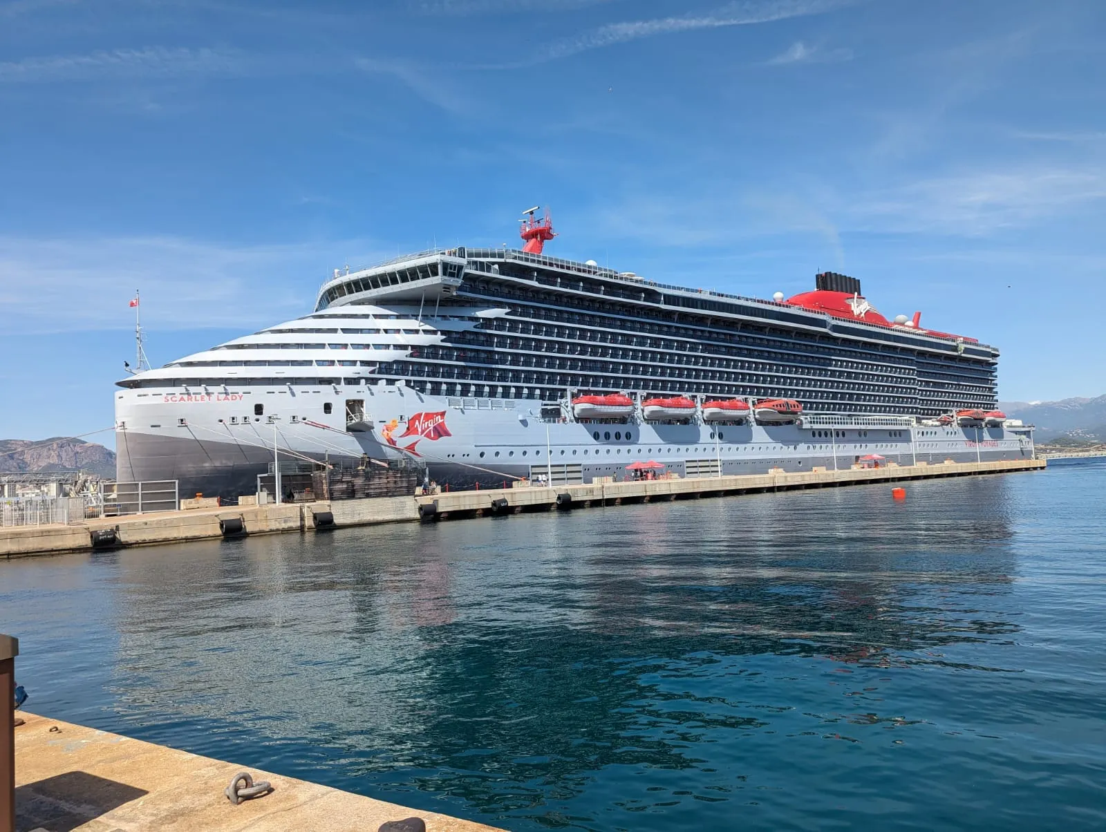 Virgin Voyages' Scarlet Lady alongside the quay on a clear day, red and white hull with lifeboats along her side and mountains behind her