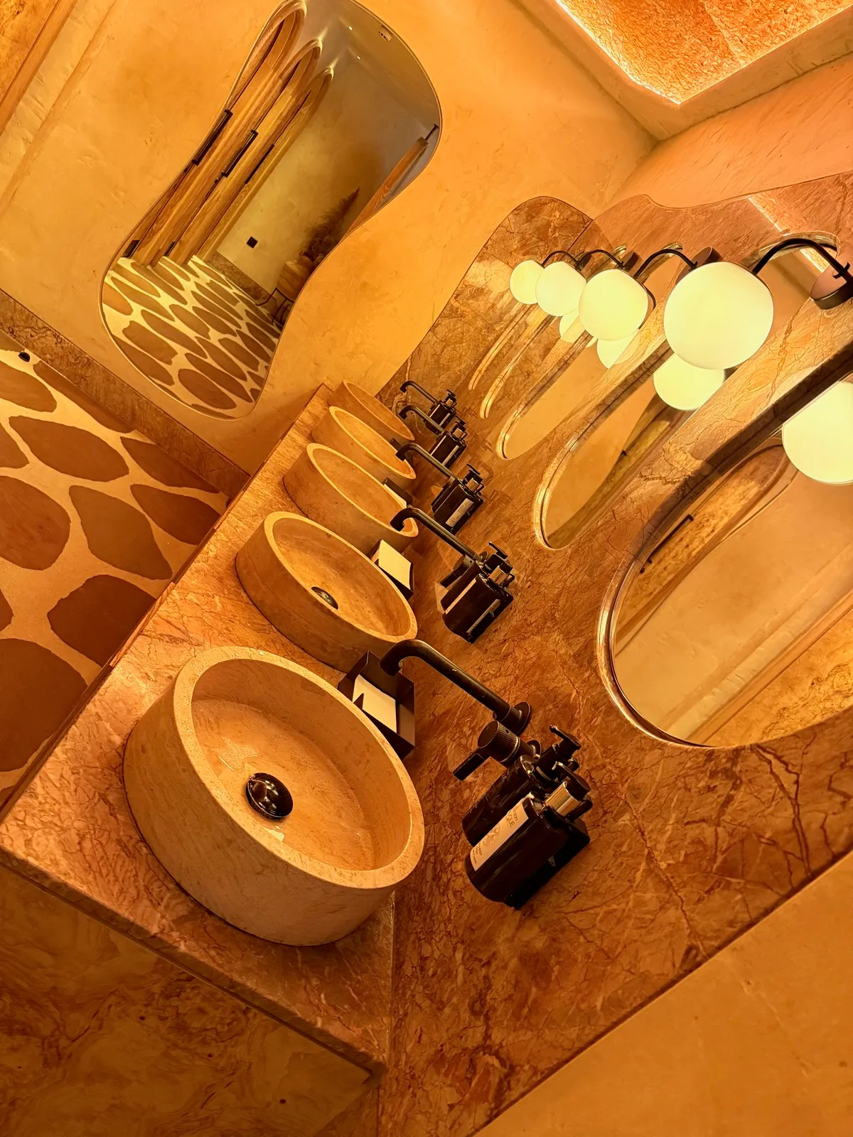 The bathroom at Fenix Manchester, with round stone basins on a travertine counter, wavy mirrors and glowing orb lights.