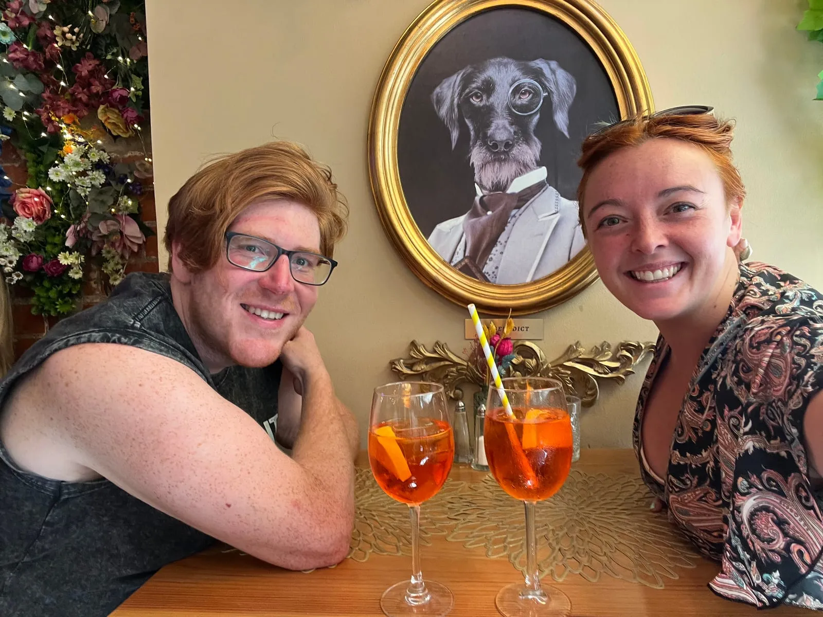 The two of us at a table in The Florentine, Barcelona, with two Aperol Spritzes in front of us and a framed portrait of a dog in a monocle and cravat on the wall behind