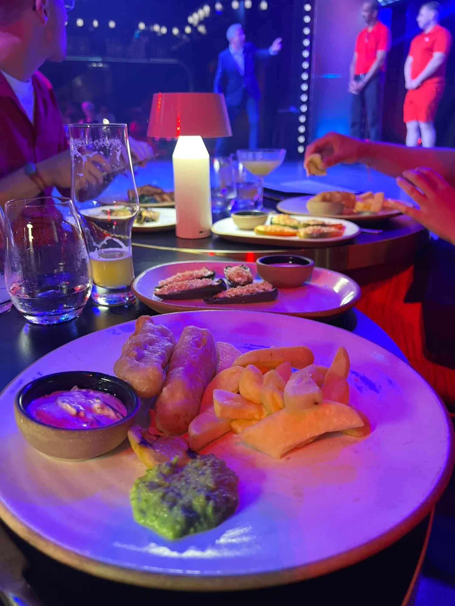 Battered fish and chips with mushy peas and a dip, plus small plates on the table, with a comedian on stage lit up behind.