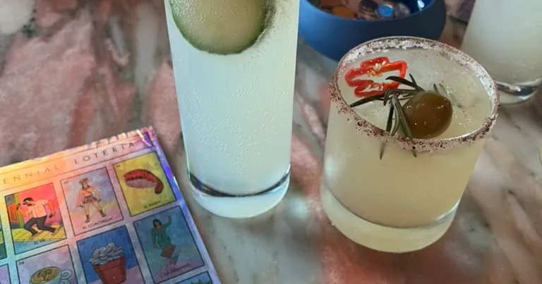 Two margaritas with salt rims on a marble table beside a holographic Millennial Lotería card