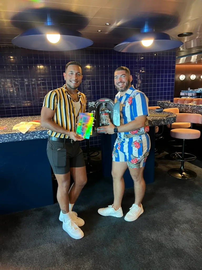The two Lotería Gigante hosts standing in the bar, one holding a Millennial Lotería card and the other a bottle of Espolòn tequila.