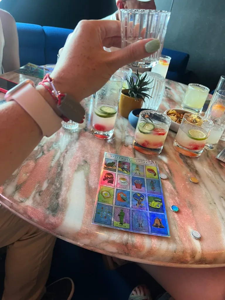 A marble table covered in margaritas, shot glasses and a bowl of snacks, with a Millennial Lotería card and counters laid out ready to play.