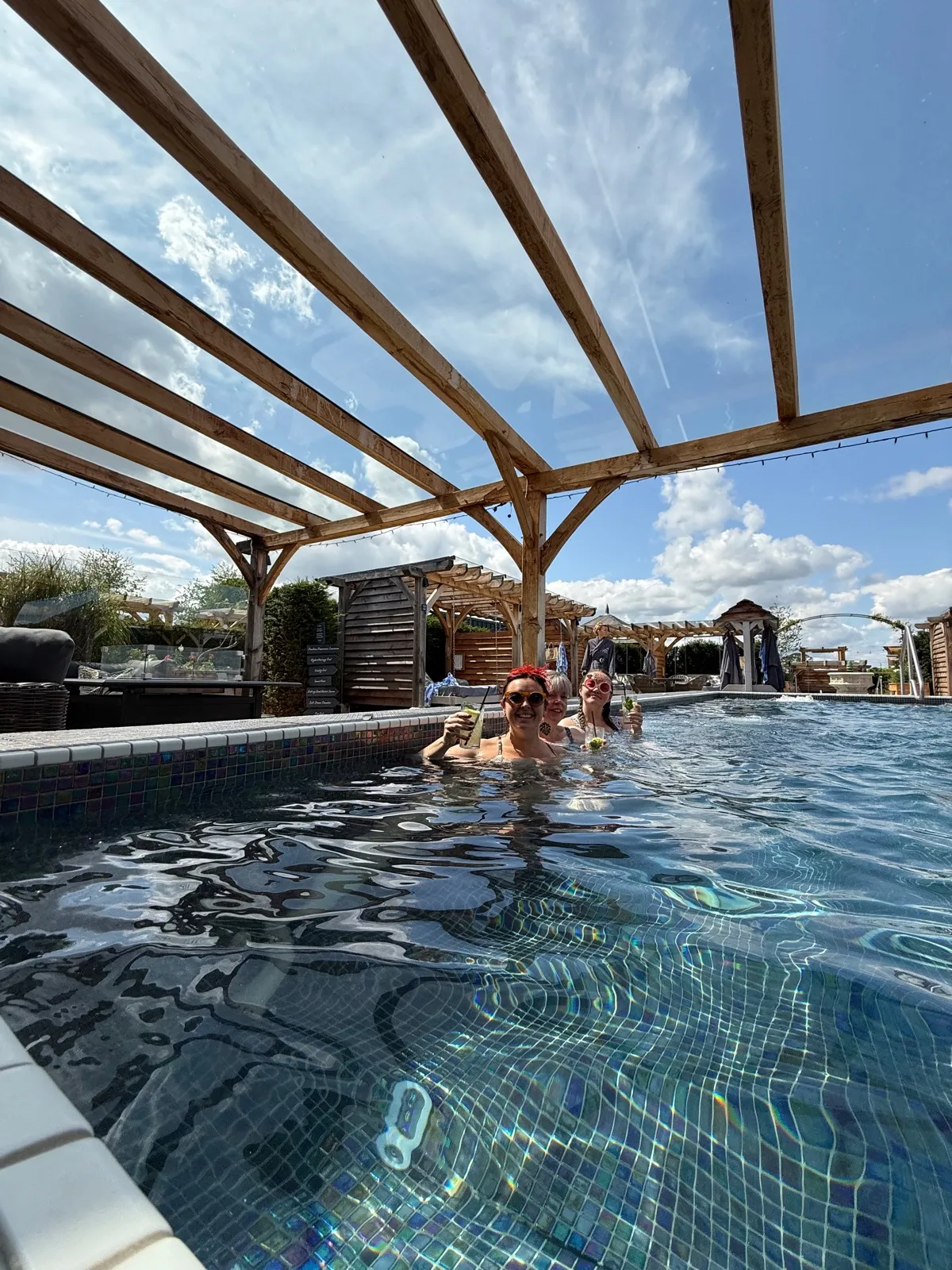 Three of us in the outdoor pool at The Manor House Alsager, drinks in hand, under a wooden pergola with the rest of the spa garden behind