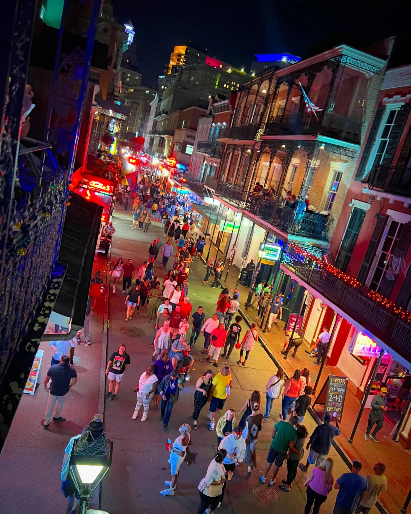 Bourbon Street at night seen from a balcony, packed with people, neon bar signs down both sides and wrought iron balconies above.