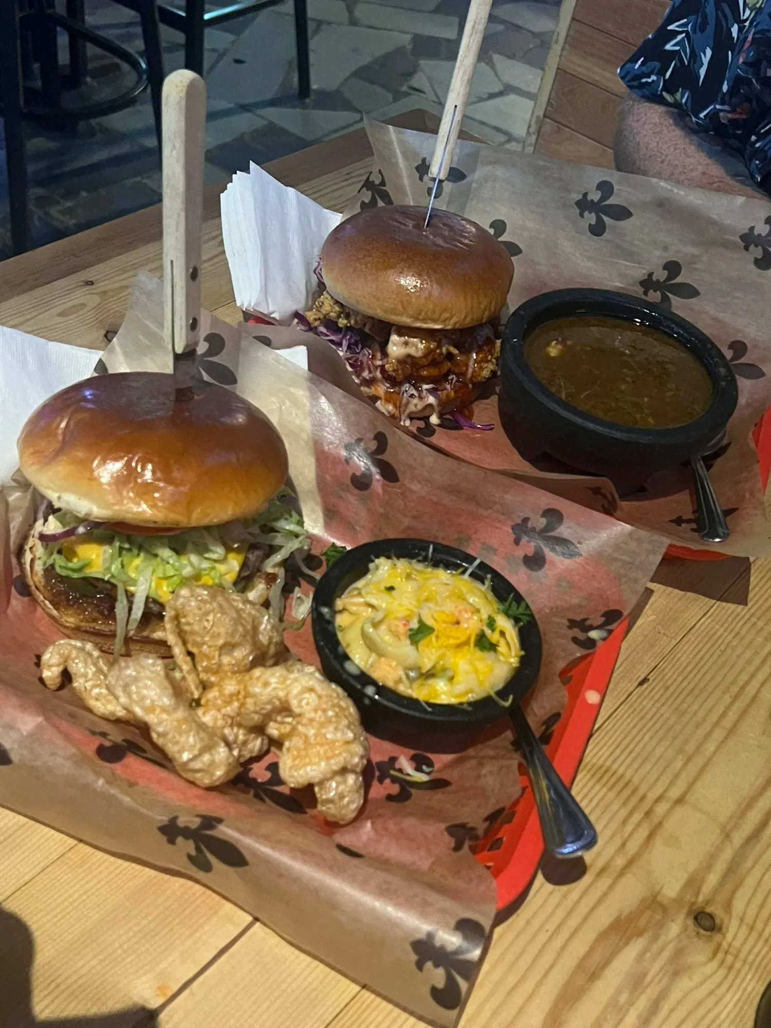 Two brioche burgers held together with skewers on fleur-de-lis printed paper, with a bowl of gumbo and a bowl of cheesy mac and cheese alongside.