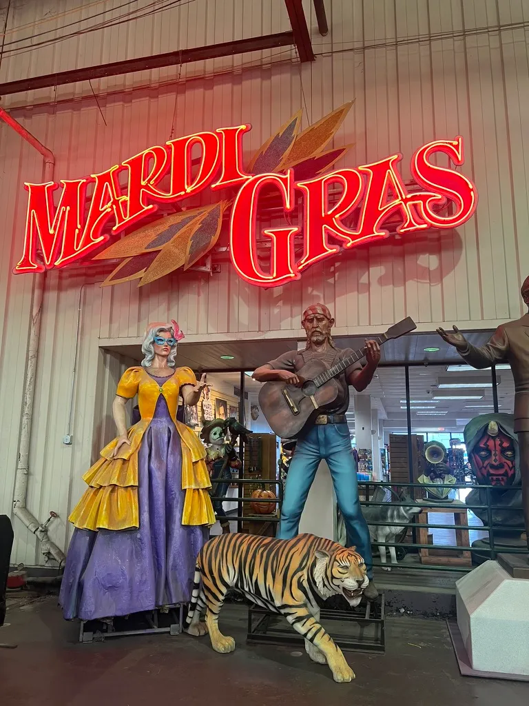Inside Mardi Gras World: a giant red neon "Mardi Gras" sign above painted parade figures, including a woman in a yellow and purple ballgown, a guitarist and a tiger.