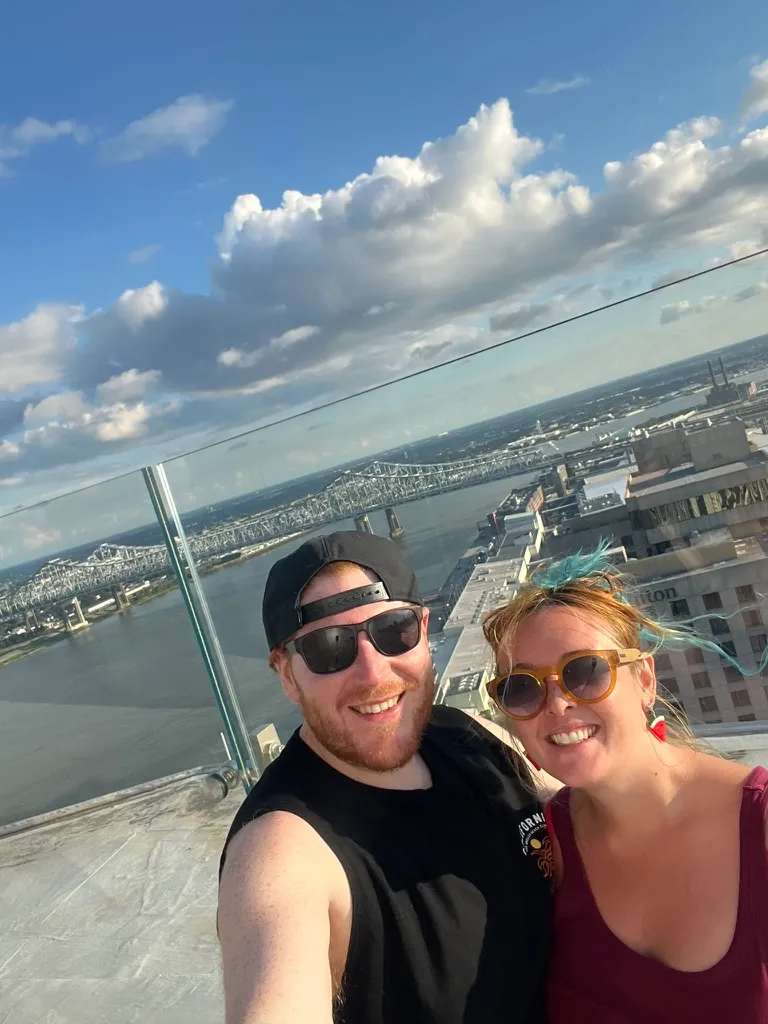 The two of us on a rooftop in New Orleans, with the Mississippi river, a long bridge and the city rooftops spread out behind us under a big cloudy sky.