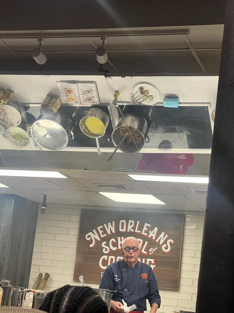 The chef at the New Orleans School of Cooking, in a navy chef's jacket and headset mic, standing in front of the painted School of Cooking sign with pans of food reflected in the mirror above him.