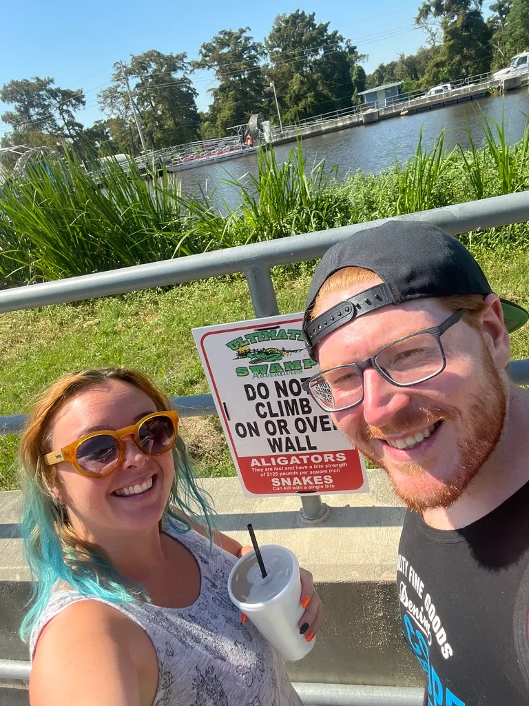 Us in front of an Ultimate Swamp Adventures sign reading "do not climb on or over wall", warning about alligators and snakes, with the launch dock and water behind.