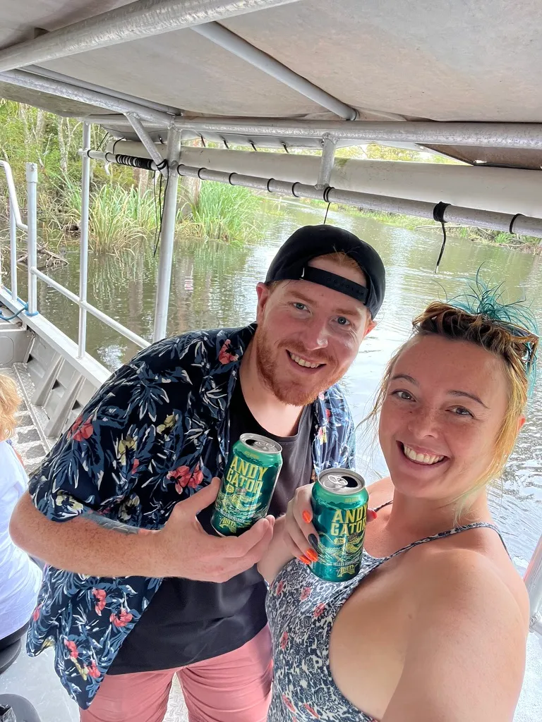 The two of us grinning on the covered deck of a swamp tour boat, holding cans of Andygator beer, with the bayou and reeds behind us.