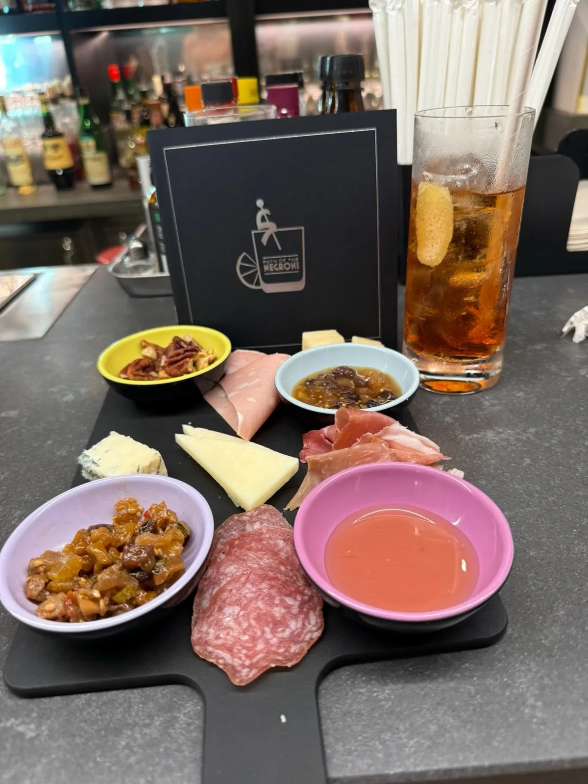 A charcuterie board of cured meats, cheeses, honey and chutney next to the Path of the Negroni booklet