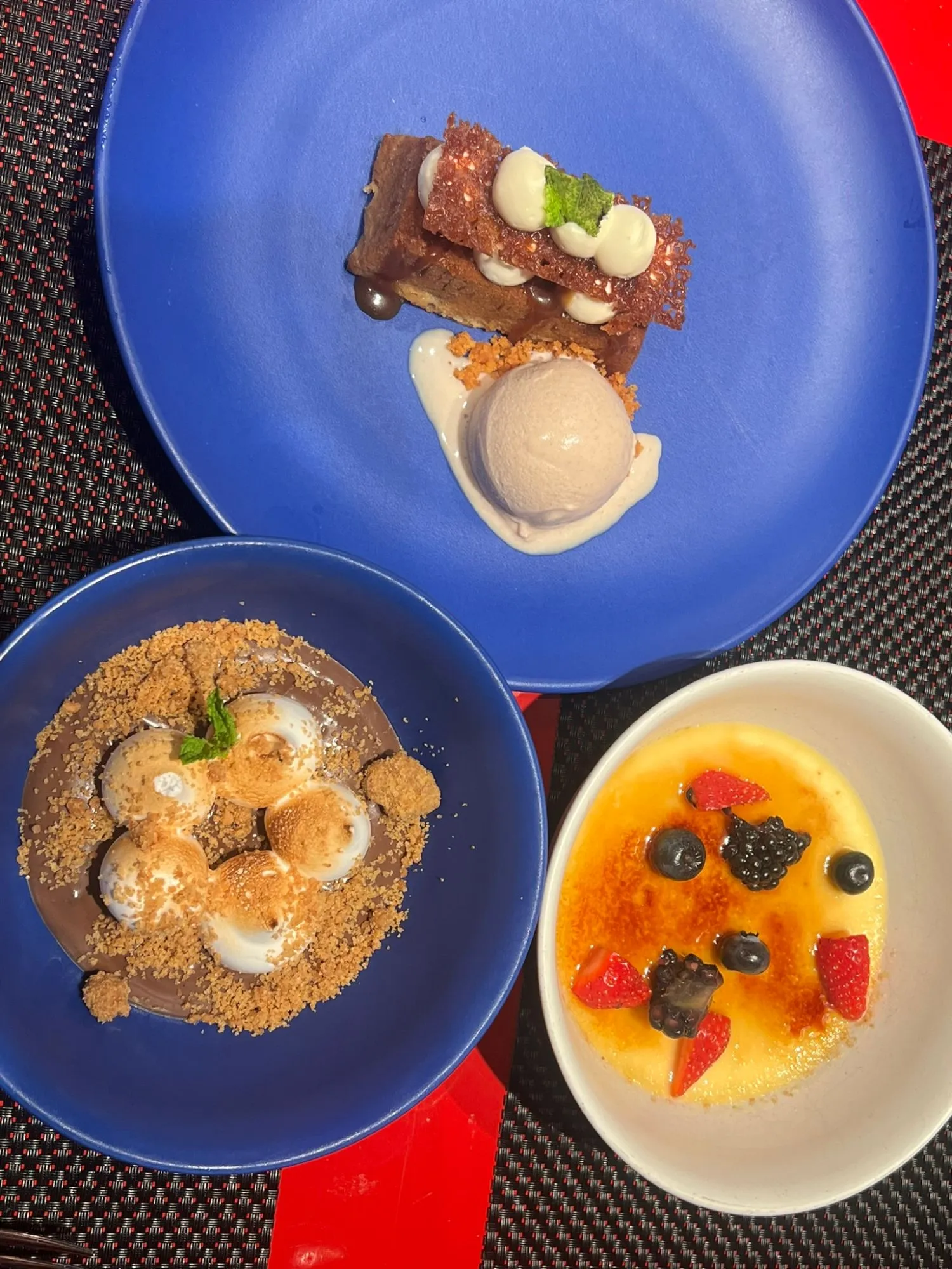Three desserts on blue plates: rum cake with a scoop of ice cream, a S'mores bowl of chocolate under toasted marshmallows and crumb, and a creme brulee topped with strawberries, blueberries and blackberries.